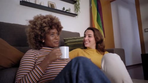 Smiling Women Couple Chatting on Sofa with LGBTQ+ Flag
