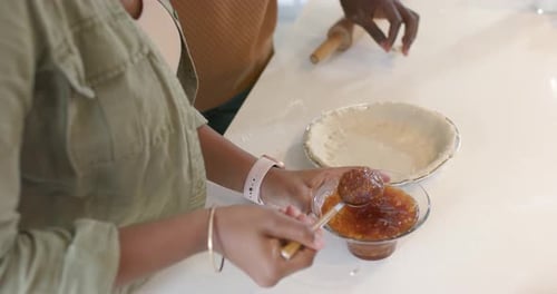 African American couple adjusting rolling pin and scooping jam into crust on countertop to bake pie