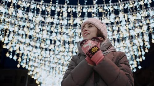 Winter Portrait of a Young Woman in a Coat and Hat Against the Background of Christmas Lights