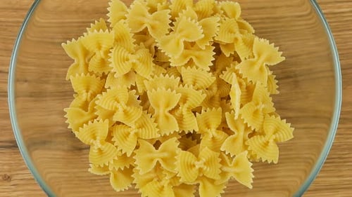 Bow-shaped pasta is poured into a glass transparent bowl against an oak wooden board. Top view.