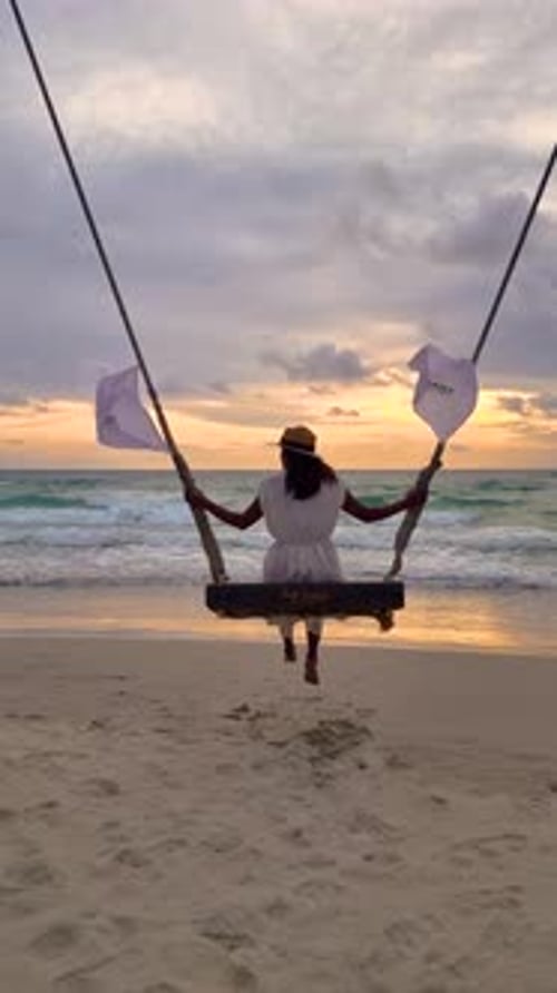 Asian Woman Watching Sunset on the Beach at a Swing Under a Palm Tree