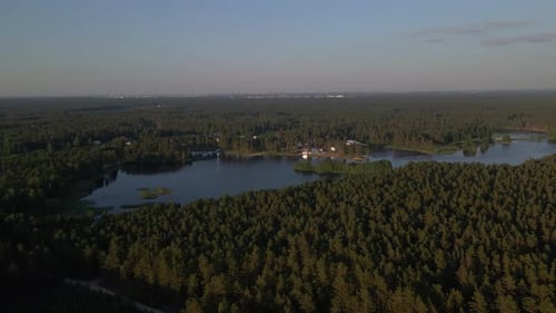 Drone orbits over a large forested lake flying to the right. Dense trees surround the lake under a c