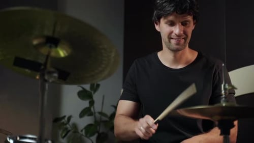 A Man in a Black Shirt Smiles While Playing Hihat with a Drumstick He is Sitting Behind a Drum Kit