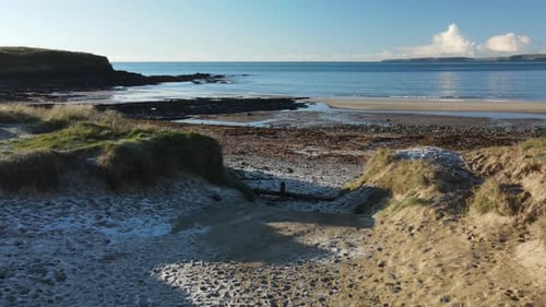 Low flying drone over frozen sand on dunes and seaweed covered beach on a calm morning with blue wat