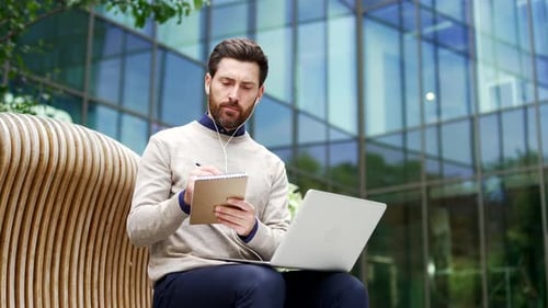 Male professional sitting outside modern building, engaged in online meeting or training. Man