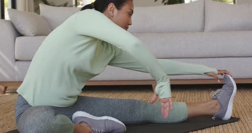 woman grasping sneaker and sliding hands along shin for hamstring stretch on mat by sofa window