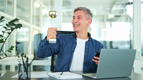 Successful Businessman Celebrates Victory with Trophy and Smartphone in Office