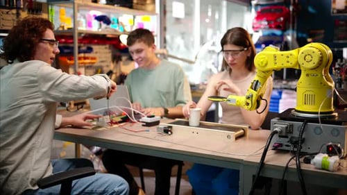 Young happy engineers programming an yellow robotic arm in the workshop to grab cardboard water glas