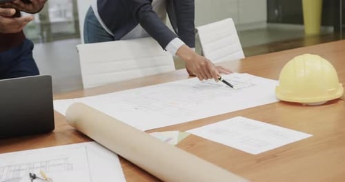 Diverse architects inspecting blueprints on table with helmet in office, in slow motion