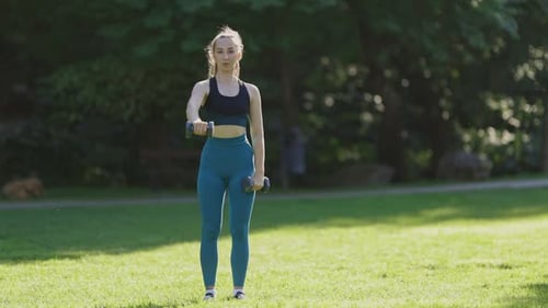 A Woman is Exercising in a Park with a Pair of Dumbbells