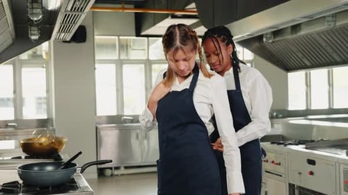 Girlfriend Chef Helps Woman Friend Tie Apron in a Restaurant Kitchen Two Adults in Uniform Prepare