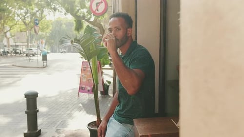Young man drinking coffee while sitting in street cafe on cityscape background