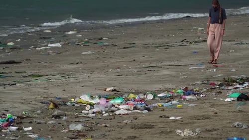 A woman walking on the beach filled with garbage washed up on the sand, static shot