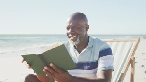 Happy senior african american man sitting on deck chair and reading book at beach, in slow motion