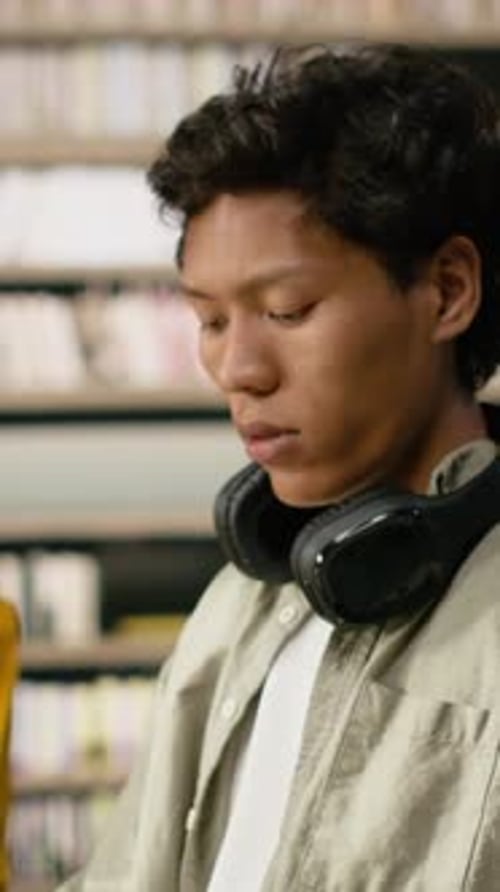 Vertical Video Student Reading Open Book at Library Desk Near Chatbot on Computer Screen