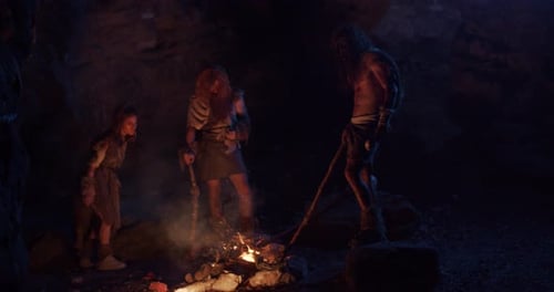 Des hommes des cavernes de la tribu, des Néandertaliens vêtus d'une peau d'animal et tenant une arme, se tiennent près du feu la nuit dans une grotte