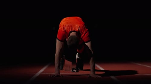 Muscular Runner Standing On Starting Blocks In Sprinter Lane Frontal View In Dark Stadium
