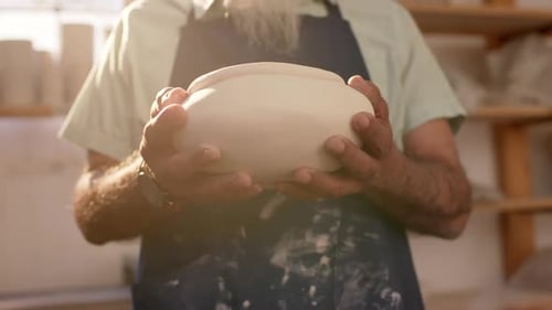 Mid section of biracial potter with long beard holding bowl in pottery studio, slow motion
