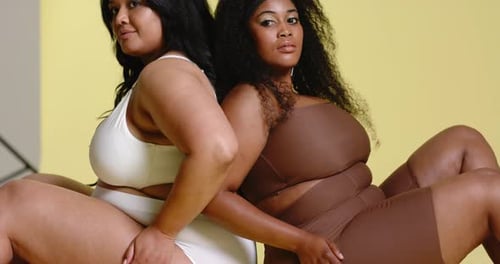 Diversas compañeras entran al set sentadas espalda con espalda en el banco junto a un fondo amarillo con trípode