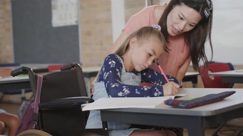 Diverse female teacher teaching schoolgirl in wheelchair writing in classroom at elementary school