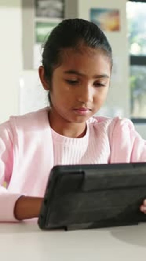 Vertical video: Leaning tween girl adjusting black tablet on school table, studying in pink sweater