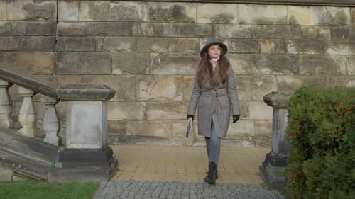 Elegant woman enjoys a peaceful walk along a stone wall in a historic park.