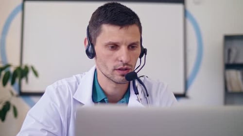 Freundlicher männlicher Arzt im weißen medizinischen Kittel mit Kopfhörern, der Telefonkonferenz auf LaptopRemote abhält