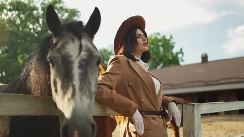 A Young Girl in a Hat Stands on a Ranch with a Horse