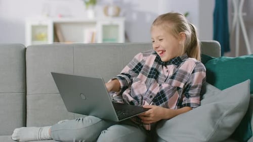 Süßes kleines Mädchen, das auf einem Sofa ruht, benutzt den Laptop, schaut sich lustige Videos und Comics an.