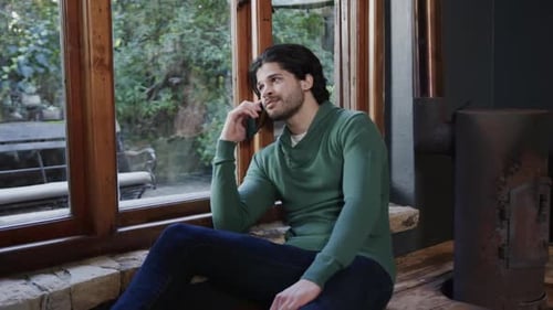 Happy caucasian man talking on smartphone sitting by window at home, slow motion