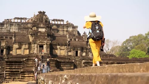 Young woman explores Cambodia's ancient temple complex wearing yellow playsuit and hat