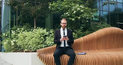 Handsome bearded man using typing mobile phone sitting on bench on city street. Attractive businessm