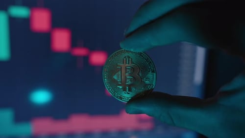 Zoomer. Main tenant une pièce Bitcoin sur fond flou de chandeliers rouges sur
un graphique en baisse
