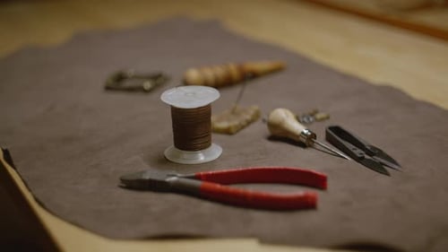 Leather crafting tools including waxed thread, awl, snips, and pliers on brown fabric