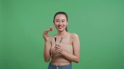 Beautiful Asian Woman Holding Makeup Brushes And Rouging Her Cheeks While Smiling On Green Screen
