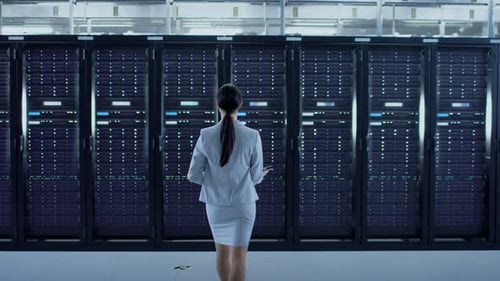Female Data Center IT Technician Walking Through Server Rack Corridor with a Laptop Computer. She i