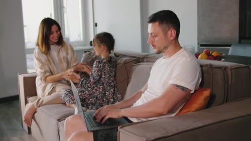 A Mother and Daughter Play on the Couch While the Father Works on His Laptop Nearby Blending Family