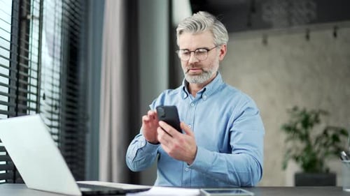 Mature gray haired bearded businessman browsing smartphone sitting at workplace in business office.