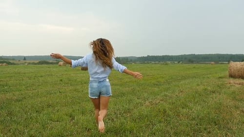 Happy Young Woman, Running On Grass Field With Haystacks. Pretty Girl In Casual Clothes Gets Fun