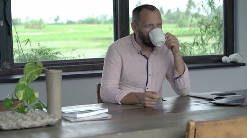 Man Sits at Table Enjoying Coffee, Laptop Nearby