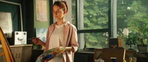 View of Young Woman Artist Painting Picture on Canvas Hold Brush and Palette Look at Camera in Light