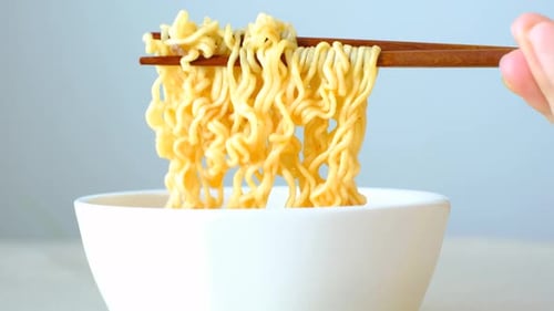 Female hand holding wooden chopsticks with instant noodles on gray background close-up.