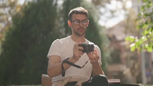 Young Man Reviews Photos on His Camera While Sitting on a Park Bench on a Sunny Day