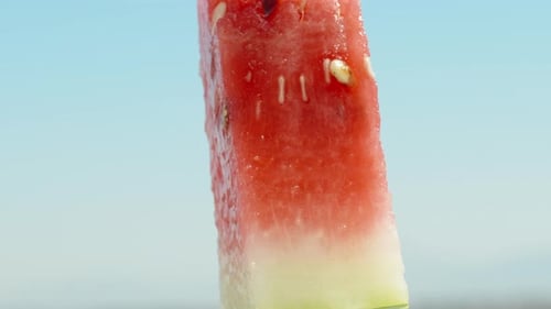 Ein Stück rote Wassermelone, das sich gegen den blauen Himmel dreht. Der Saft fließt ihn hinunter. Nahaufnahme.