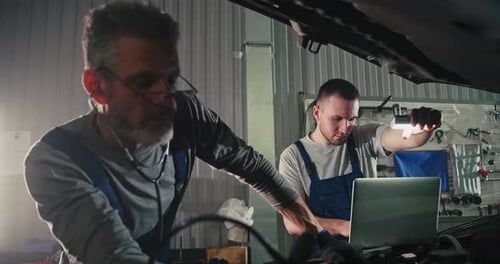 Two Mechanic Caucasian Man in Large Garage Inspecting Parts Under Raised Car