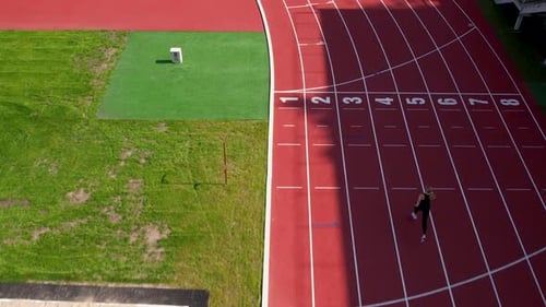 A female athlete captured from above runs down the red track past the grassy field and sandpit, main