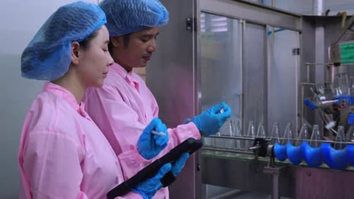Two asian factory worker inspect glass bottles on conveyor belt using digital tablet.