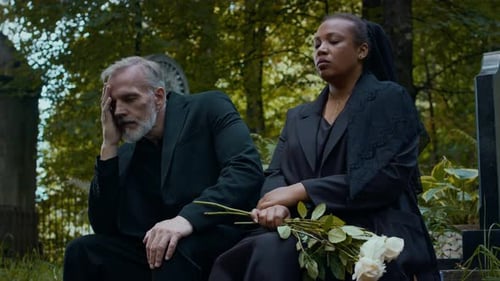 Bereaved Couple Dressed in Black Sitting with Flowers in Cemetery