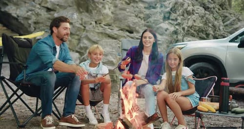 Happy smiling mother father with two children cooking sausages on bonfire at forest camp near car
