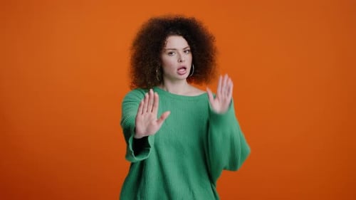 Entschlossene junge Frau in grünem Strickpullover mit STOP-Handzeichen auf orangefarbenem Hintergrund